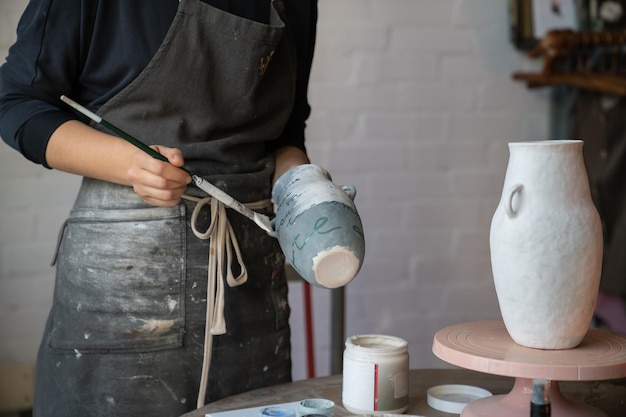 Female artisan in apron creates ceramic tableware painting white vase with brush in workshop