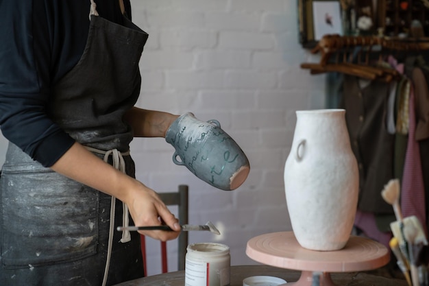 Female artisan in apron creates ceramic tableware painting white vase with brush in workshop