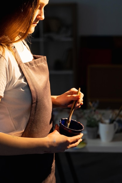 Female artist Home art Creative hobby Inspiration process Calm smiling woman in apron holding bowl with blue paint mixing brush in light flash interior room