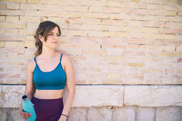 Female athlete rests leaning against a brick wall holding a bottle of water healthy lifestyle concept