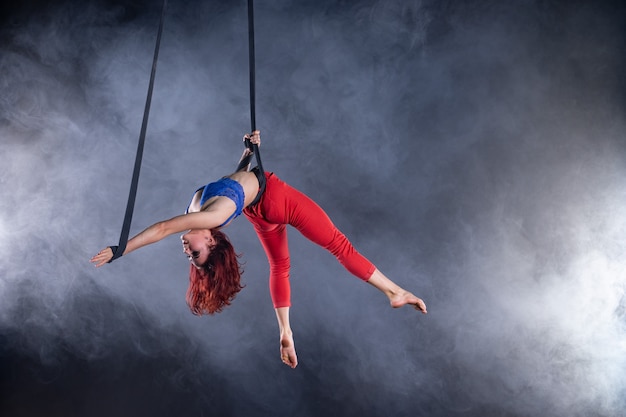 Female athletic, sexy and flexible aerial circus artist with redhead on aerial straps on black.