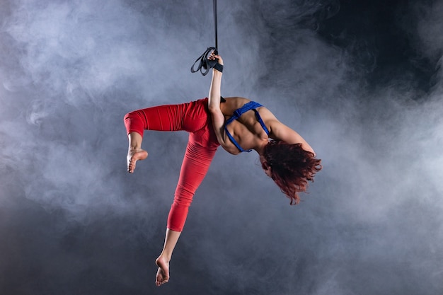 Female athletic, sexy and flexible aerial circus artist with redhead on aerial straps on black.