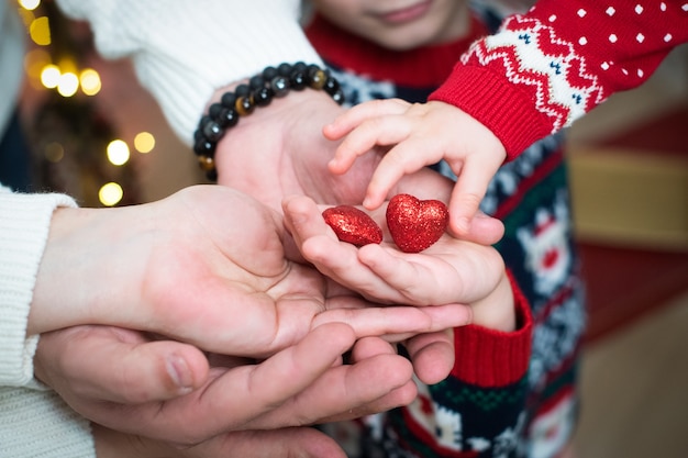 Female, baby and male hands are holding two red, shiny hearts. Valentine's day, love, support, trust.