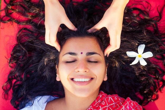 Female beautician doctor with patient make procedure to beautiful indian girl.close up portrait of asian woman closed eyes and white smile wellness head massaging on beach.Face lifting massage in spa