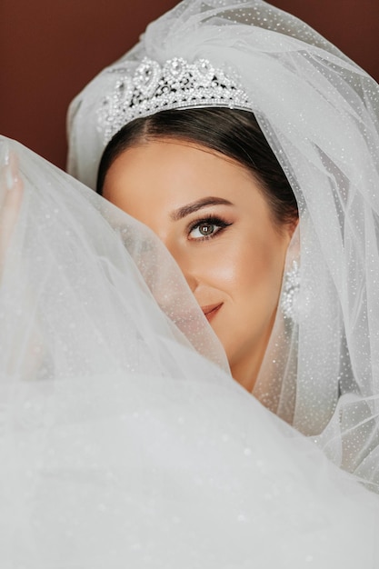 Female beauty Cute woman at home The bride poses for the photographer A stylish woman wears a white dress Professional makeup and hair The bride has a crown on her head and a long veil