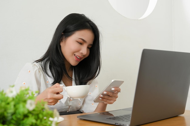 Female business entrepreneur sitting at her office workspace sipping coffee and using her smartphone