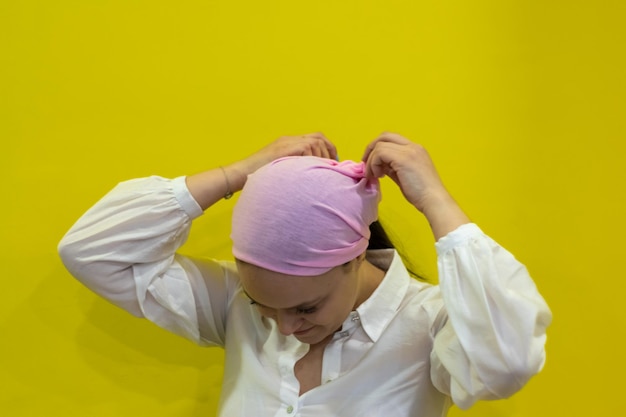 Female cancer patient putting on a pink headscarf on yellow background Concept of fighting and beating cancer