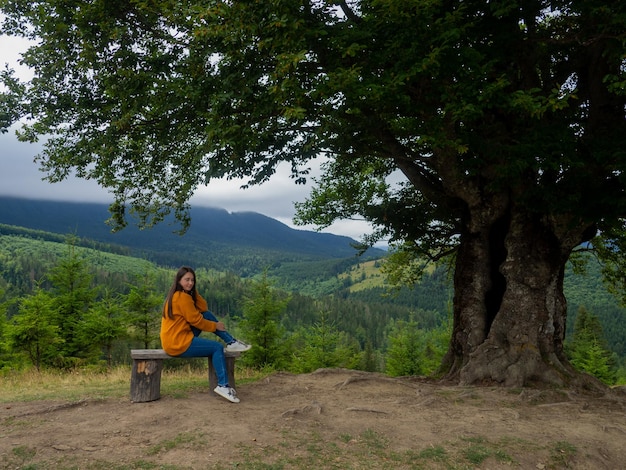 Female in casual clothes sits under large tree with forest background