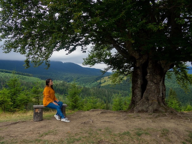 Female in casual clothes sits under large tree with forest background