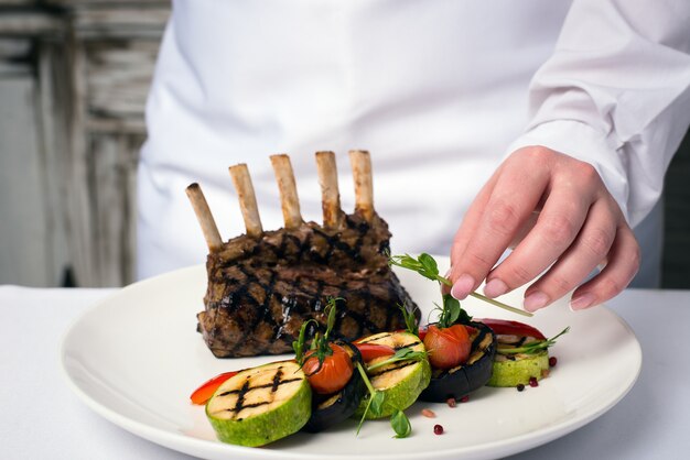 Female chef in the kitchen of a hotel or restaurant, only hands, she finishes a vegetable dish with meat on a white plate.