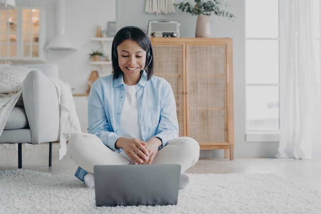 Female coach in headset conducts online consultation on laptop by video call sitting on the floor