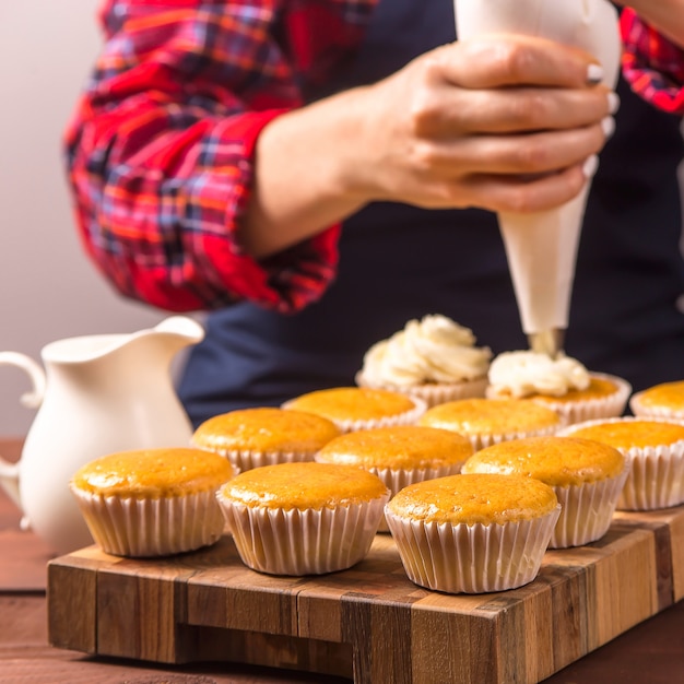 Female confectioner in a blue apron and a plaid red shirt applies cream to cupcakes 