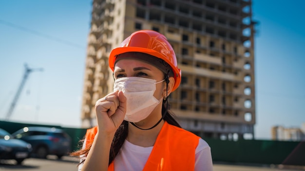 Female construction worker in overalls and medical mask coughing into fist on background of house under construction Concept of threat of coronavirus epidemic infection