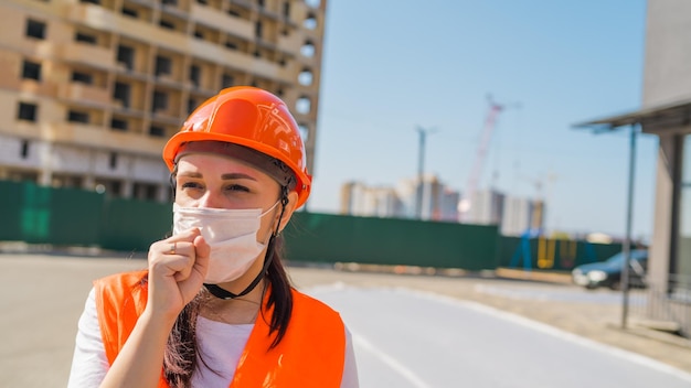 Female construction worker in overalls and medical mask coughing into fist on background of house under construction Concept of threat of coronavirus epidemic infection