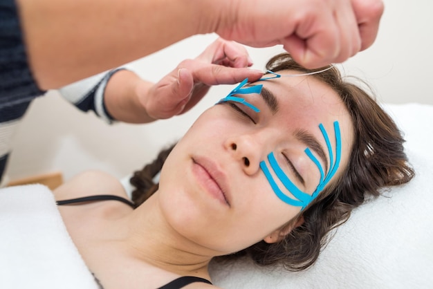 Female cosmetician doing the taping of the face of the patient lying on the desk in the officeSpecialist performing kinesio taping application on girl's face