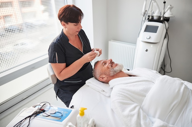 Female cosmetologist holding a cotton swab while an elderly man lying on a spa bed