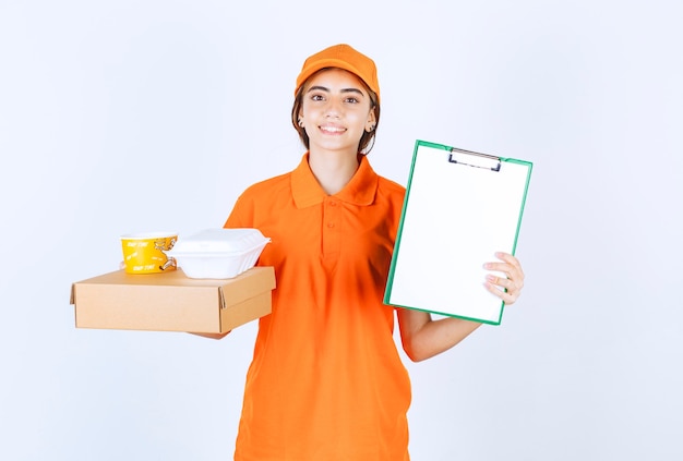 Female courier in orange uniform holding yellow and white takeaway boxes, cardboard parcel and a customer list for signature