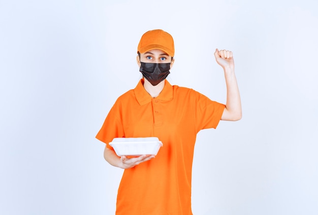 Female courier in yellow uniform and black mask holding a plastic takeaway food box and showing her fist.