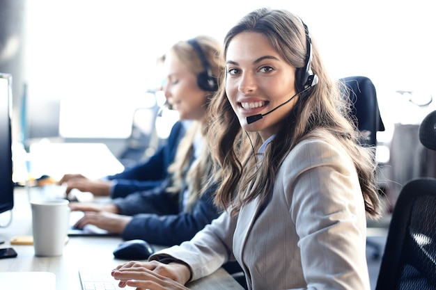 Female customer support operator with headset and smiling, with collegues at background.