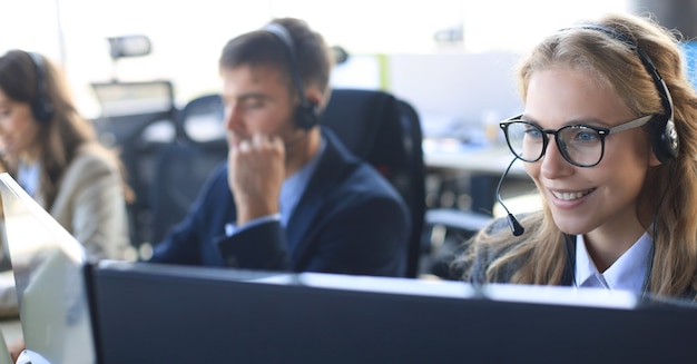 Photo female customer support operator with headset and smiling, with collegues at background.