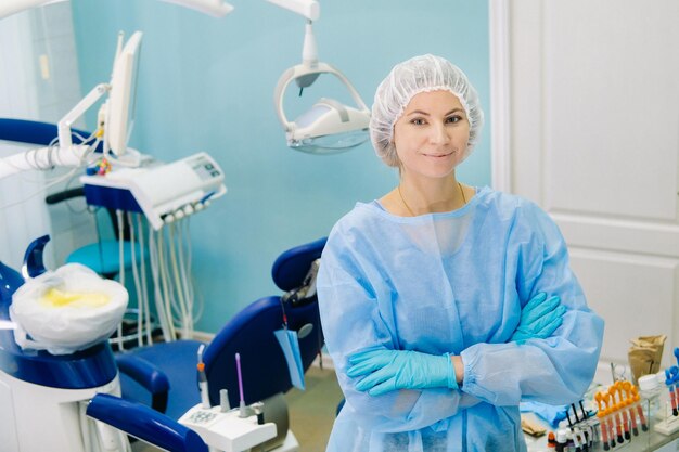 A female dentist wearing a medical mask and rubber gloves poses for the camera and folds her arms in her office