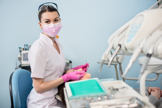 Female dentist with dental tools - mirror and probe treating patient teeth and looking to the camera at dental clinic office.