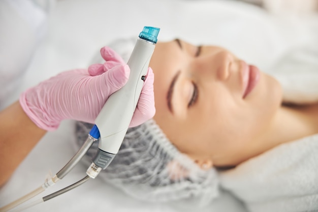 Female dermatologist holding a beauty device in her hand over her young patient with closed eyes