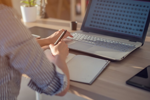 Female designer using laptop, sketching at blank notepad. Woman hand writing in notebook on wooden desk.