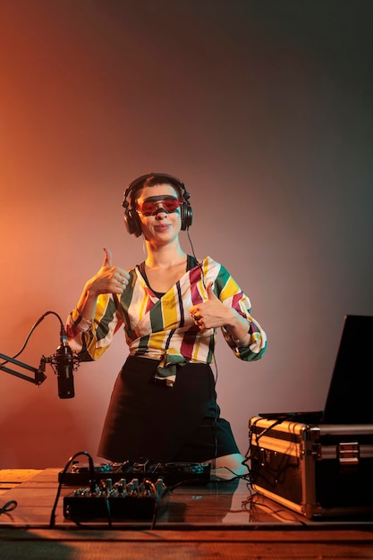 Female DJ doing thumbs up and like sign, showing approval symbol to celebrate success and achievement. Mixing techno music with turntables and expressing positive agreement gesture.