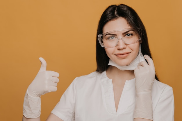 Female doctor in glasses, dressed in a medical gown, shows thumb up gesture, taking off her surgical mask.