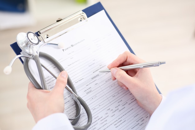 Female doctor hand holds silver pen filling patient list at clipboard pad closeup.