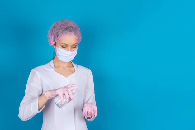 A female doctor in a medical mask, cap and gloves with an antiseptic in her hands on a blue wall. Isolate