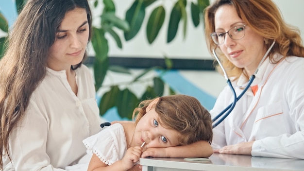 Female doctor pediatrician using stethoscope listen to the heart of happy healthy cute kid girl at m...