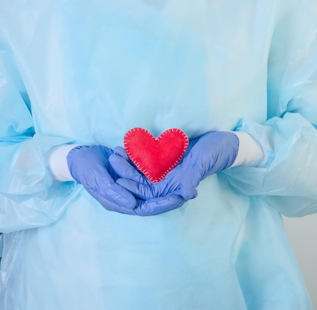 A female doctor in a protective suit holds a red heart in her hands in gloves on a white background