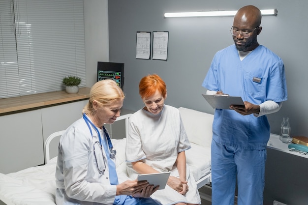 Female doctor showing something on tablet pc to senior woman while they sitting on the bed in ward with nurse making notes in medical card