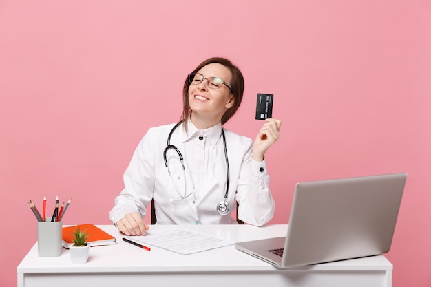 Female doctor sit at desk work on computer with medical document credit card in hospital isolated on pastel pink wall background. Woman in medical gown glasses stethoscope. Healthcare medicine concept