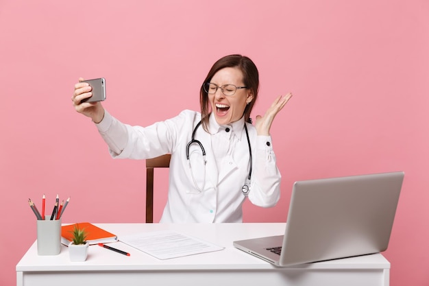 Female doctor sit at desk work on computer with medical document hold cellphone in hospital isolated on pastel pink background. Woman in medical gown glasses stethoscope. Healthcare medicine concept.