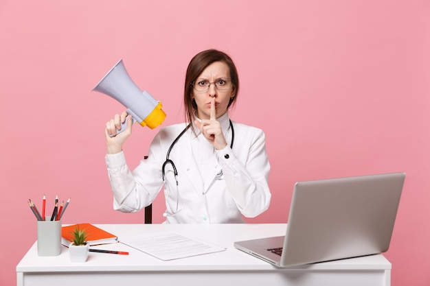 Female doctor sit at desk work on computer with medical document hold megaphone in hospital isolated on pastel pink background. Woman in medical gown glasses stethoscope. Healthcare medicine concept.