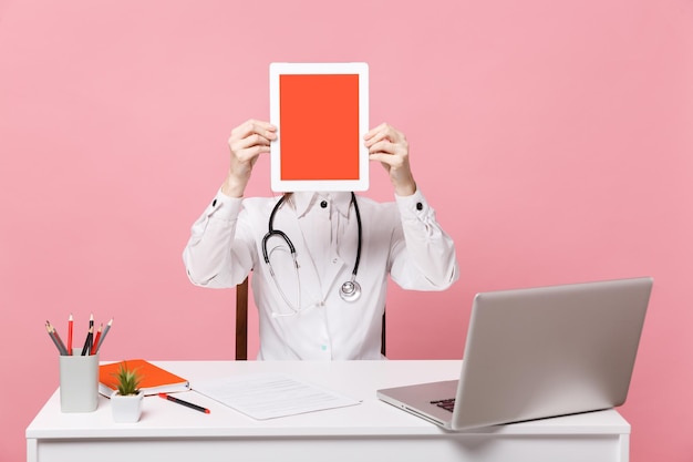 Female doctor sit at desk work on computer with medical document hold tablet in hospital isolated on pastel pink wall background. Woman in medical gown glasses stethoscope. Healthcare medicine concept