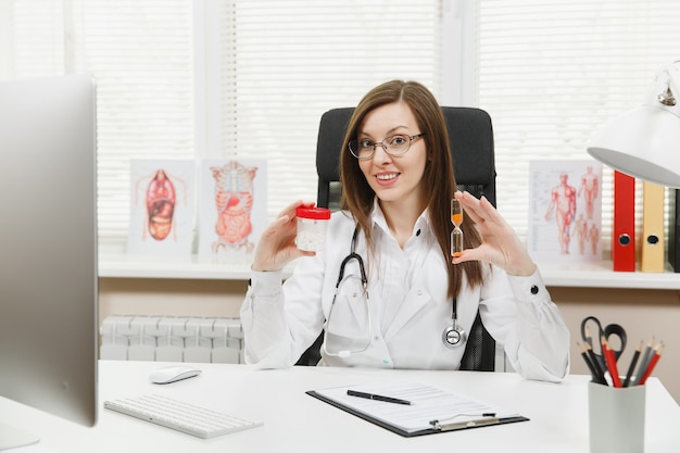 Female doctor sitting at desk, holding bottle with white pills, hourglass, working with medical documents in light office in hospital