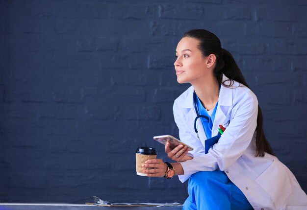 Female doctor sitting with mobile phone and drinking coffee