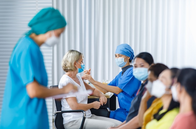 Female doctor wears face mask and hospital uniform injecting coronavirus vaccine into Caucasian senior woman shoulder while other multinational patients wait in queue in blurred foreground.