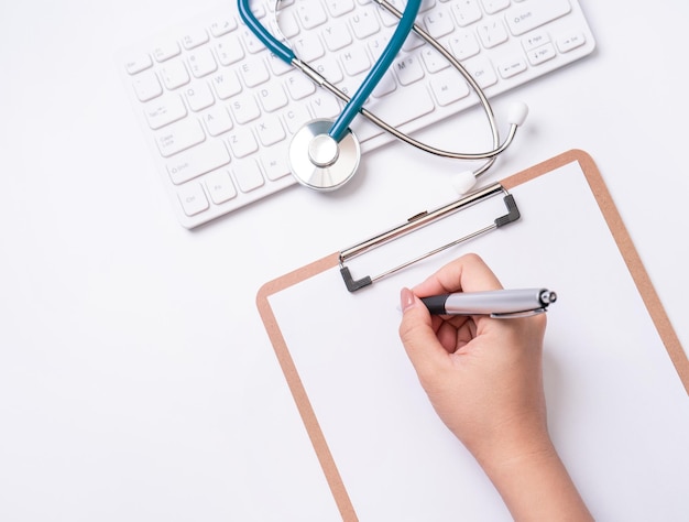 Photo female doctor writing a medical record case over clipboard on white working table with stethoscope computer keyboard top view flat lay copy space