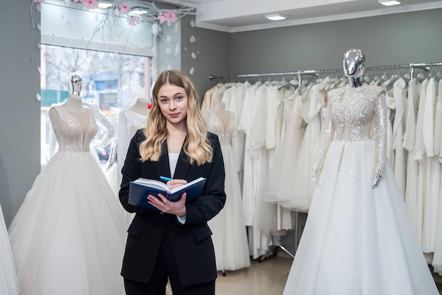 Photo female dressmaker with diary and pen posing near bridal wear in wedding boutique