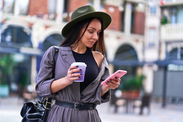 Female drinking coffee and using smartphone during walking in the center of a european city