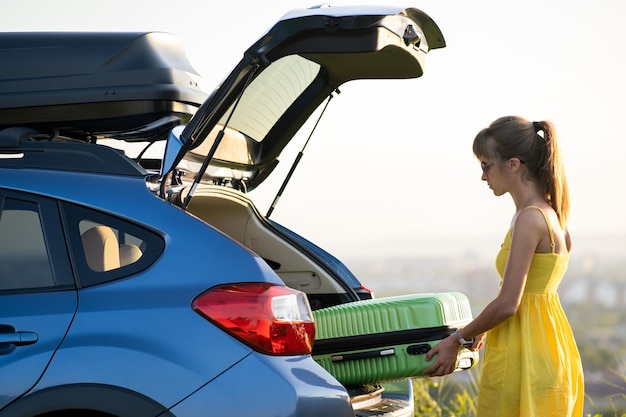 Female driver in summer dress putting green suitcase inside her car trunk Travel and vacations concept