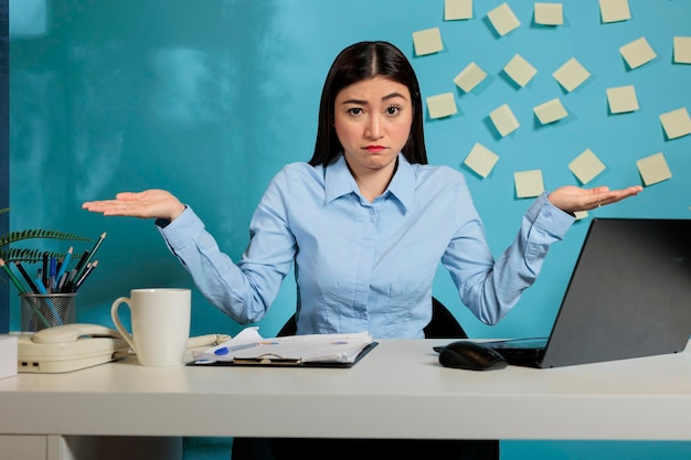 Female employee sitting in modern office making dont know gesture to camera, feeling hesitant and unclear. Insecure professional woman with no idea what should do to finish project.