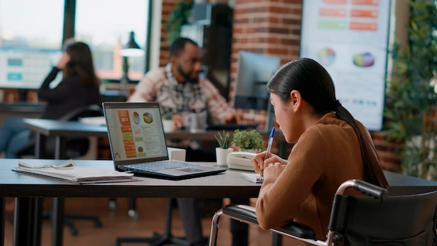 Female employee taking notes on business documents, looking at charts on laptop and working on financial statistics. Worker planning marketing growth in disability friendly office. Handheld shot.