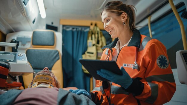 Female EMS Professional Paramedic Using Tablet Computer to Fill a Questionnaire for the Injured Patient on the Way to Hospital Emergency Care Assistant Comforting the Patient in an Ambulance