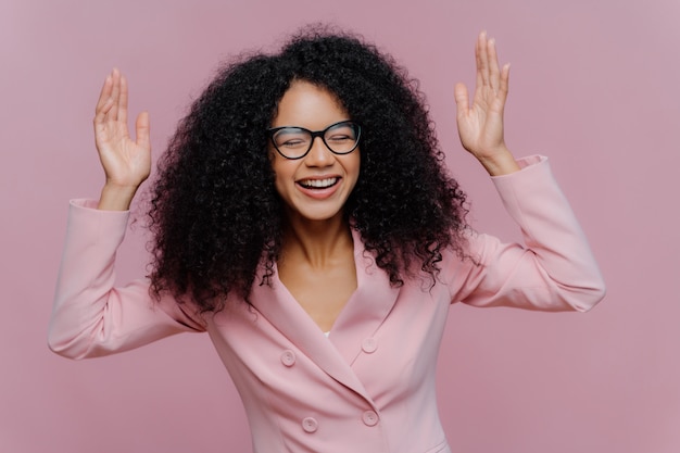 Female entrepreneur raises hands, laughs sincerely and wears spectacles and formal jacket